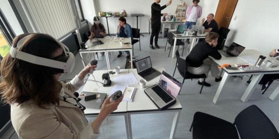 Mehrere Personen arbeiten in einem hellen Seminarraum an Tischen mit Laptops. Im Vordergrund testet eine Person mit einer Virtual-Reality-Brille und Controllern eine virtuelle Anwendung, während weitere Personen die Situation beobachten, diskutieren oder an ihren Computern arbeiten. Die Szene vermittelt einen Eindruck von kollaborativer Forschung zu virtuellen Arbeitswelten und Mensch-Technik-Interaktion.