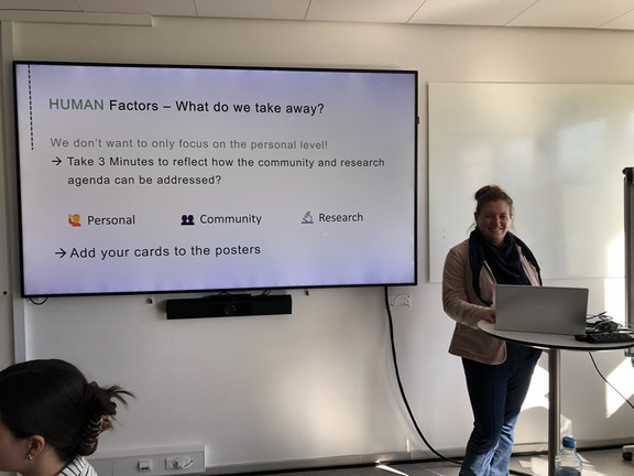 Frauke Mörike steht an einem kleinen Stehtisch mit Laptop und präsentiert in einem Seminarraum. Neben ihr hängt ein großer Bildschirm mit einer Präsentationsfolie mit dem Titel „HUMAN Factors – What do we take away?“. Auf der Folie steht unter anderem, dass nicht nur die persönliche Ebene betrachtet werden soll und dass drei Minuten Zeit zum Nachdenken über Community- und Forschungsaspekte genommen werden sollen. Außerdem sind die Punkte „Personal“, „Community“ und „Research“ aufgeführt sowie die Aufforderung, Karten zu den Postern hinzuzufügen.