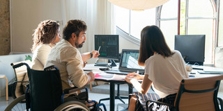 Three people, consisting of a man in a wheelchair and two women, work cooperatively on several screens.