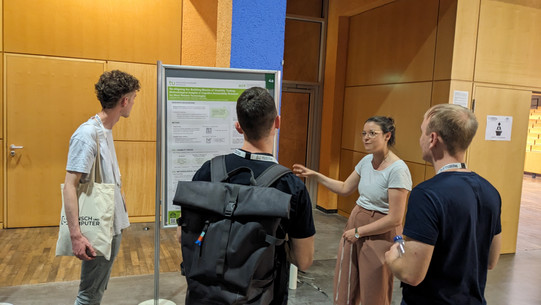 Jana-Sophie Effert points to a scientific poster entitled “Re-Aligning the building blocks for usability testing: methodological insight in cognitive accessibility research for work-related technologies.” Three people stand in front of her and look at the poster, with their backs to the camera.
