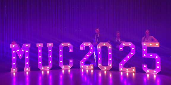  Violet illuminated letters stand out against a purple background. Behind them are Hendrik Sellmann, Frauke Mörike, and Jana-Sophie Effert leaning against the illuminated letters.