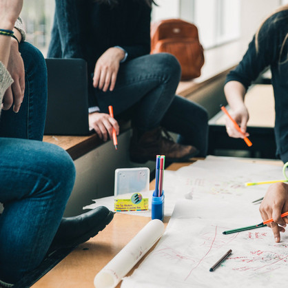 Four people are sitting at a desk writing something on a poster. There are also pencils and a cup on the table.