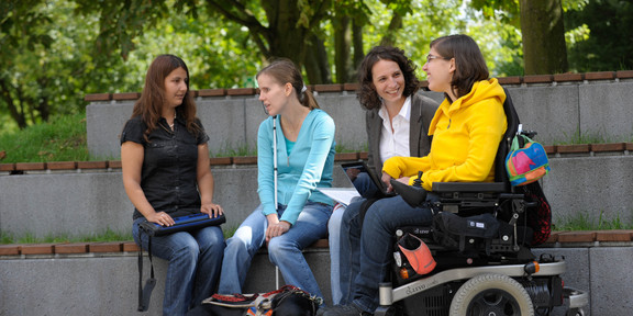 Vier Studierende, darunter Studierende mit Behinderung, sitzen gemeinsam auf dem Campus.