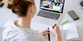 Student sitting at desk working on laptop, next to it are books and pens.